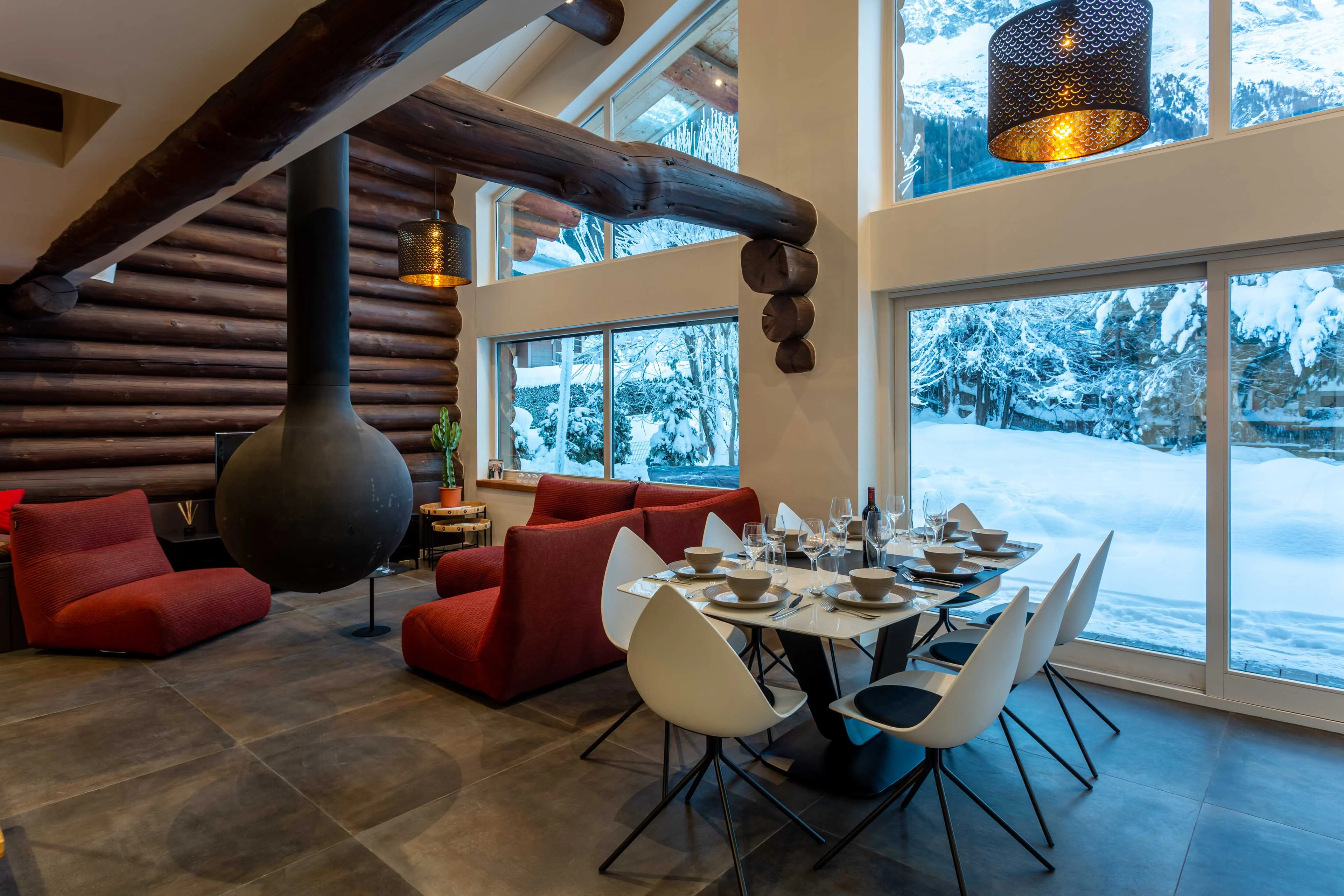 Salle à manger ouverte d'un chalet à Chamonix avec poêle à bois suspendu et baies vitrées donnant sur la neige