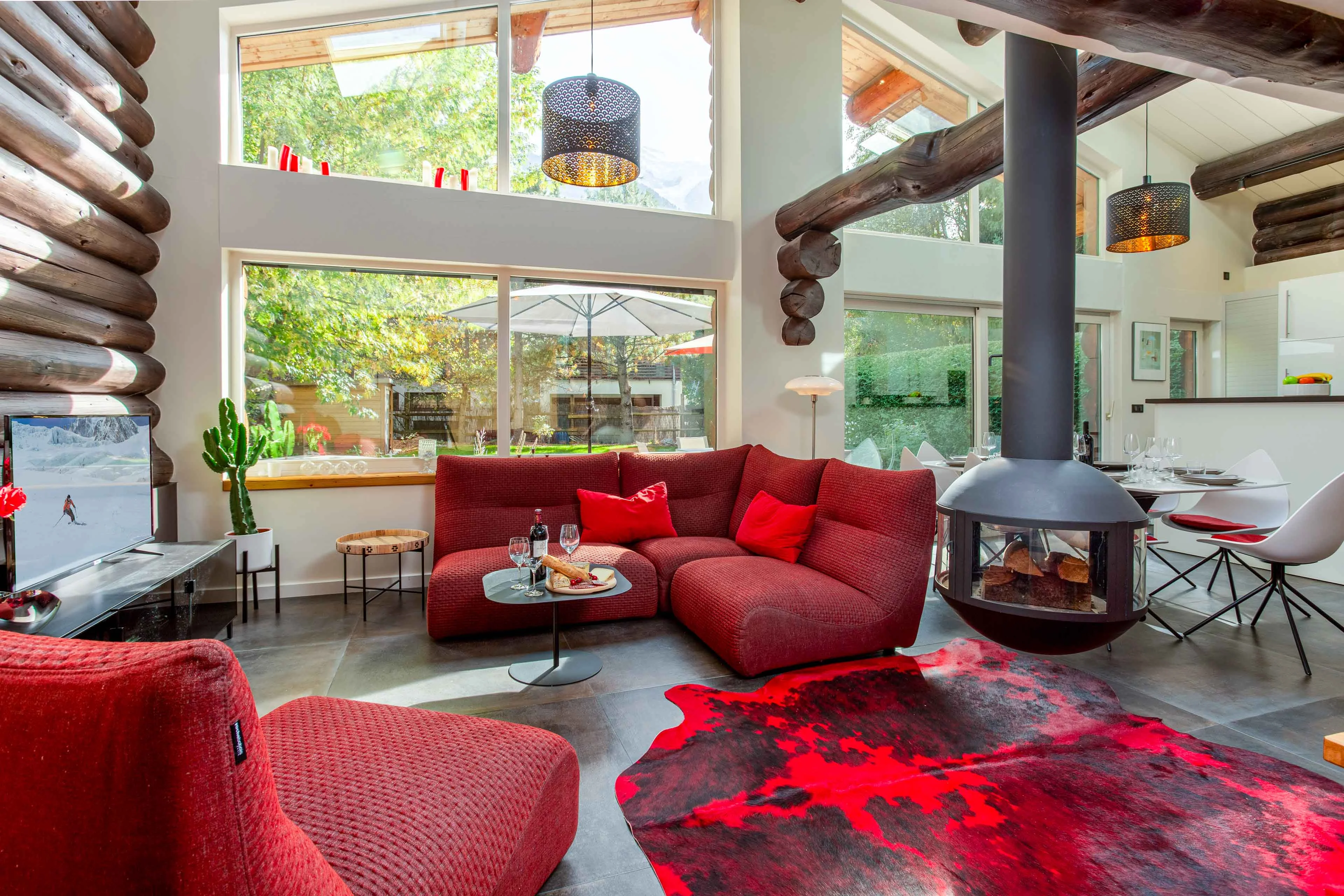 Salon de chalet à Chamonix avec poêle à bois suspendu, canapés rouges et baies vitrées donnant sur le jardin