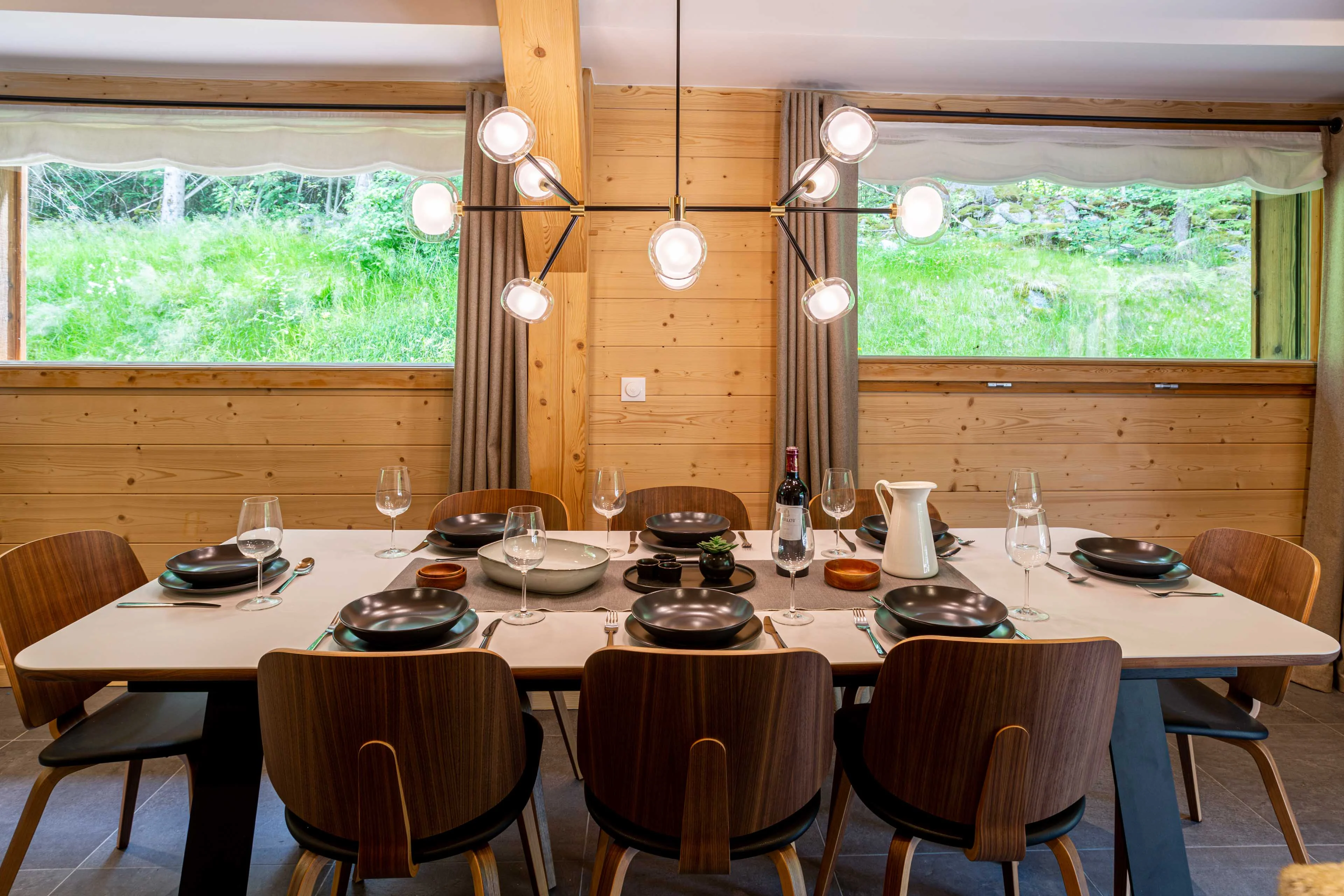 Table dressée pour huit personnes dans un chalet à Chamonix, avec lambris en bois et baies vitrées donnant sur la prairie en été