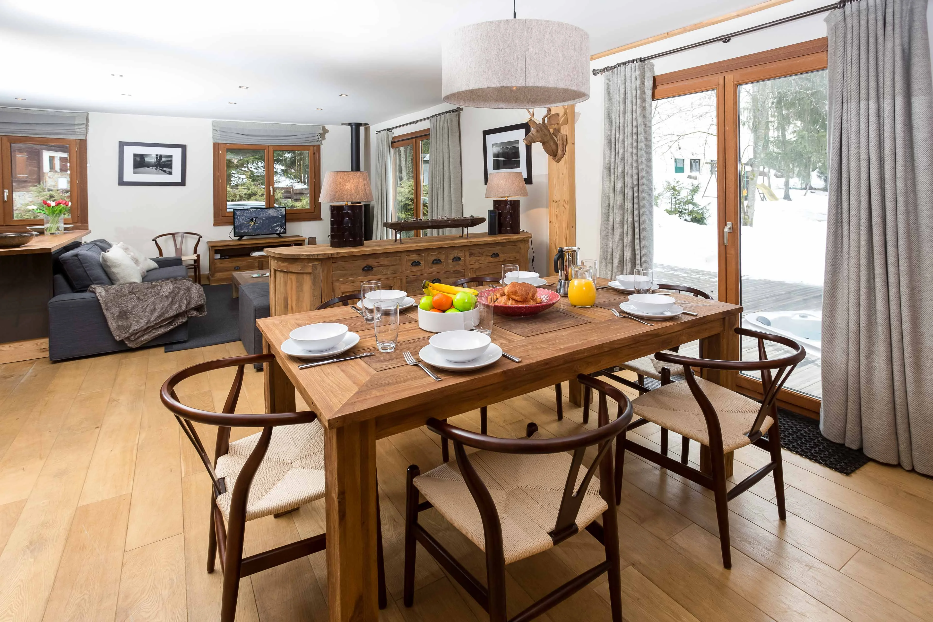 Dining table laid for breakfast in an open plan Chamonix chalet, with the living room behind and snow outside